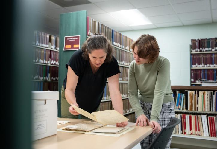 A photo of two females. On the left is a professor and a student is on the right. They are leaning over a table in an archive, looking at a document.