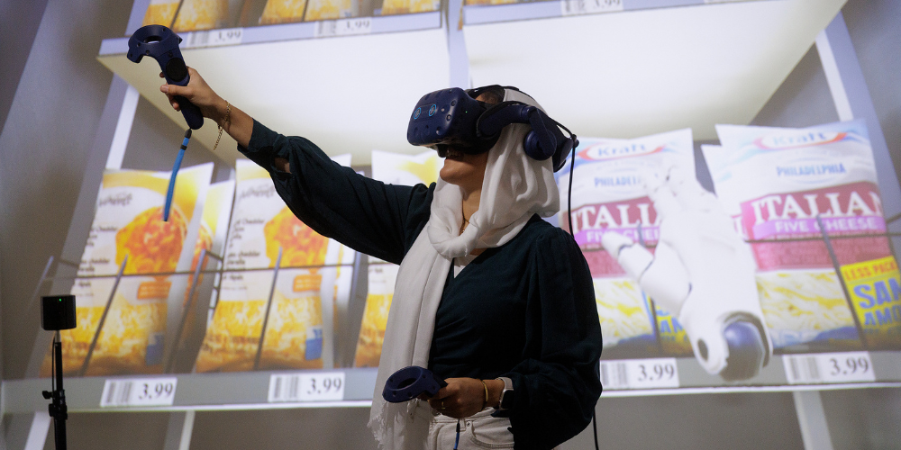 A student wearing a VR headset moves with a grocery screen behind her