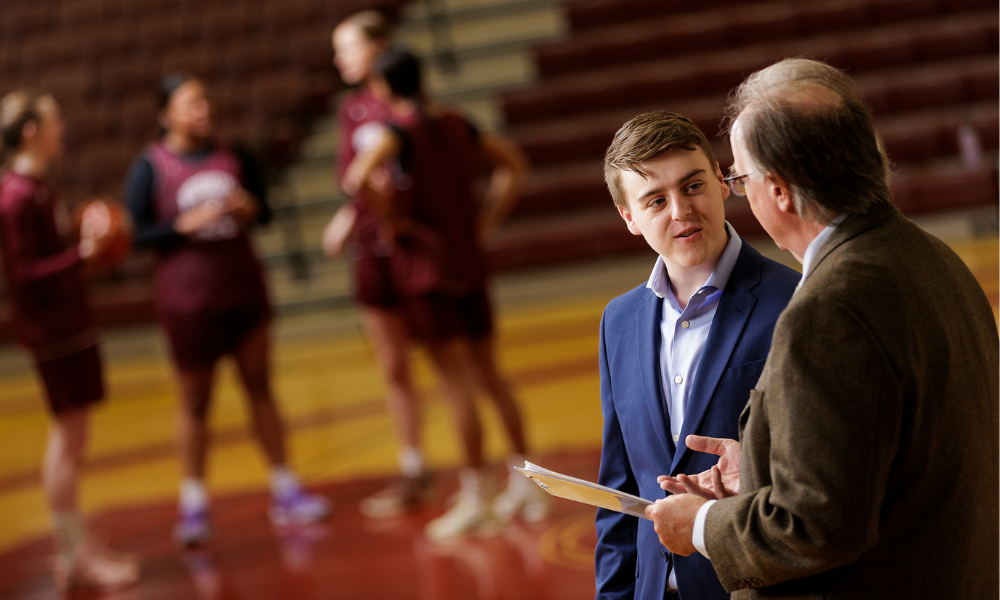 A business student talks with a professor while a basketball team practices behind them