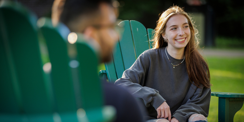 A student sits on a green chair outdoors, the back of another student's head is in the foreground