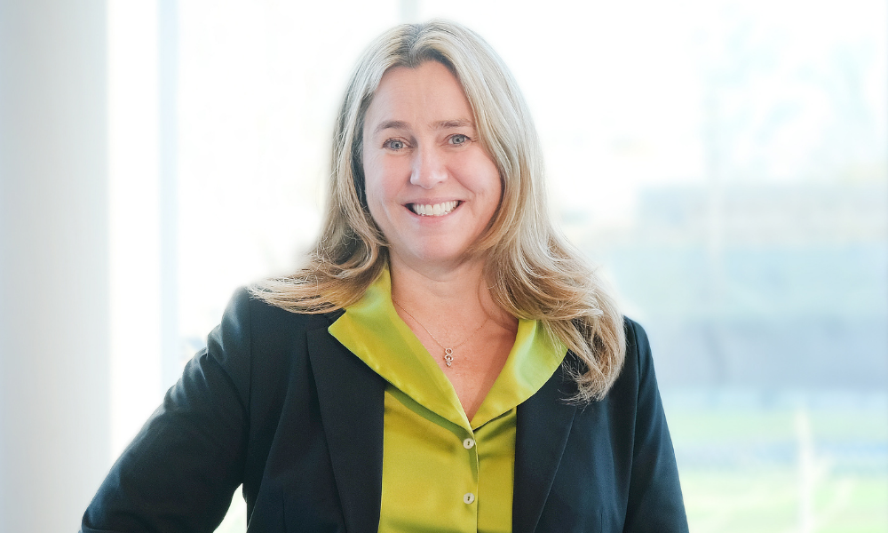 Susan wears a green shirt and black blazer. She has long blonde hair.